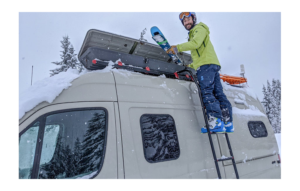 Man on ladder getting skis out of the storage on top of the Winnebago Revel.