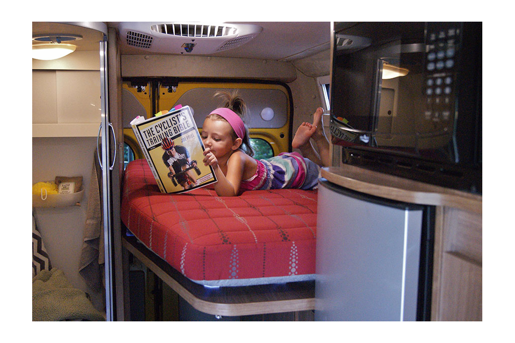 Little girl reading a book on back bed of a RV.