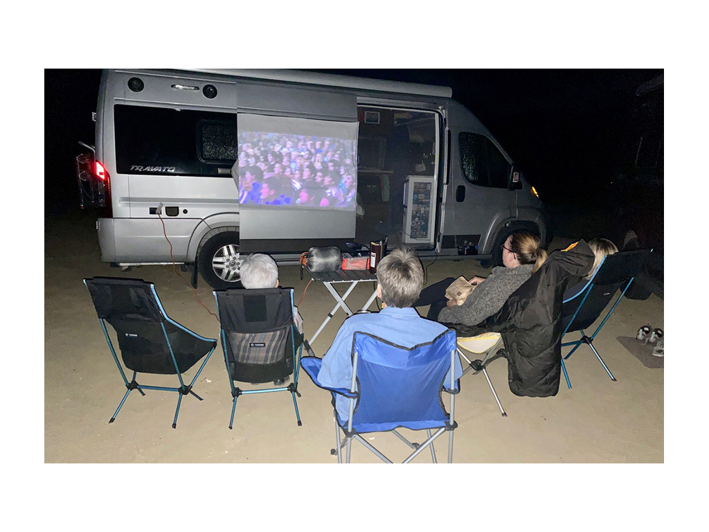 Group of people sitting in camp chairs watching a movie projected onto a sheet hanging on Travato