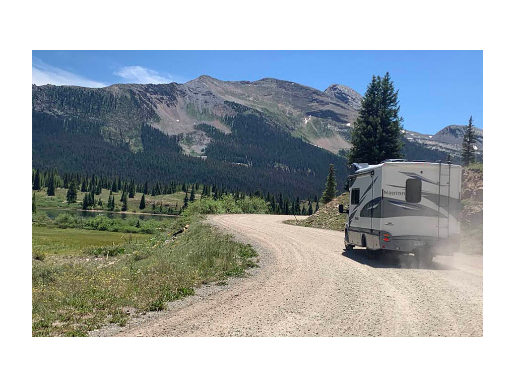 Navion driving down dirt road in Colorado. Green trees and mountains are in distance