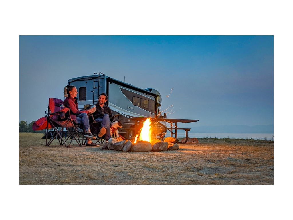 Katelyn and Howard sitting next to a campfire with a small black and white dog by their side. Their Navion is parked behind them.