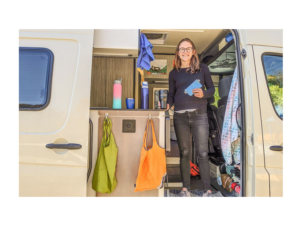 Kelly standing in Revel doorway with reusable water bottles, coffee mugs, and bags