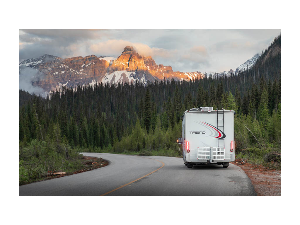 Winnebago Trend driving down tree lined road toward the snow covered mountains ahead.