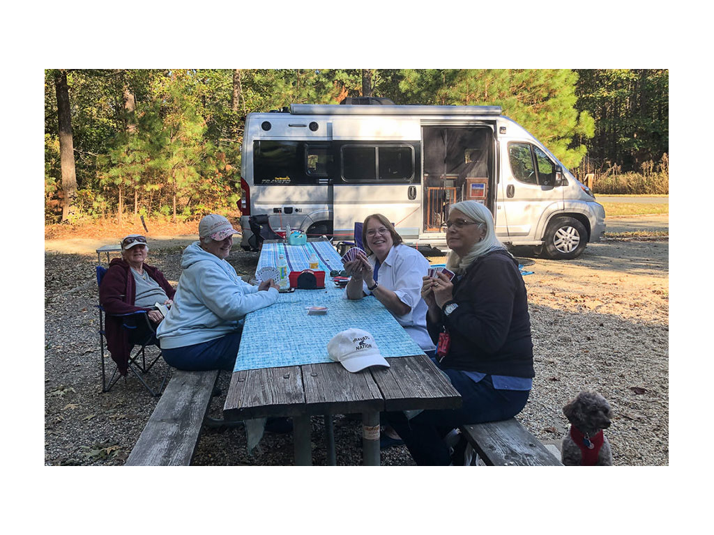 4 people sitting at a picnic table playing cards with a Travato in the background.