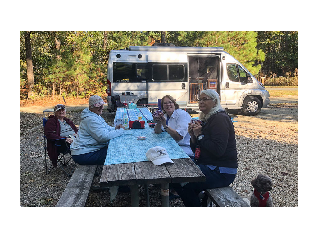 4 people sitting at a picnic table playing cards with a Travato in the background. 