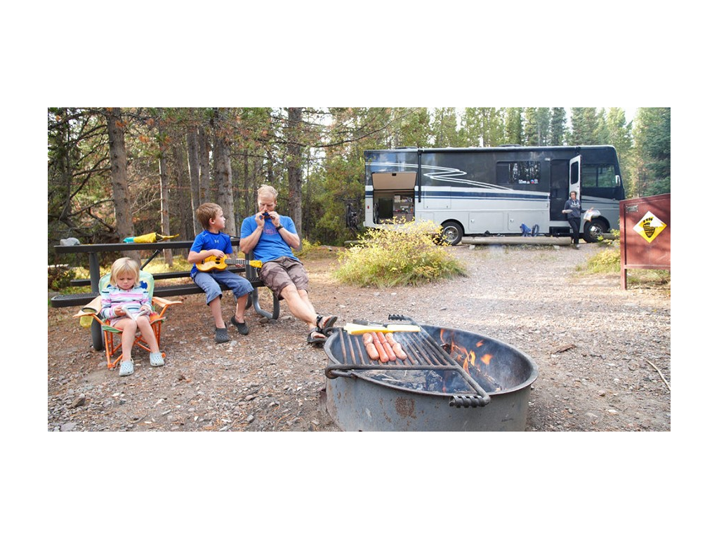 Eli and kids sitting at picnic table next to fire pit with hotdogs roasting. Kelly is stepping out of Adventurer carrying supplies.
