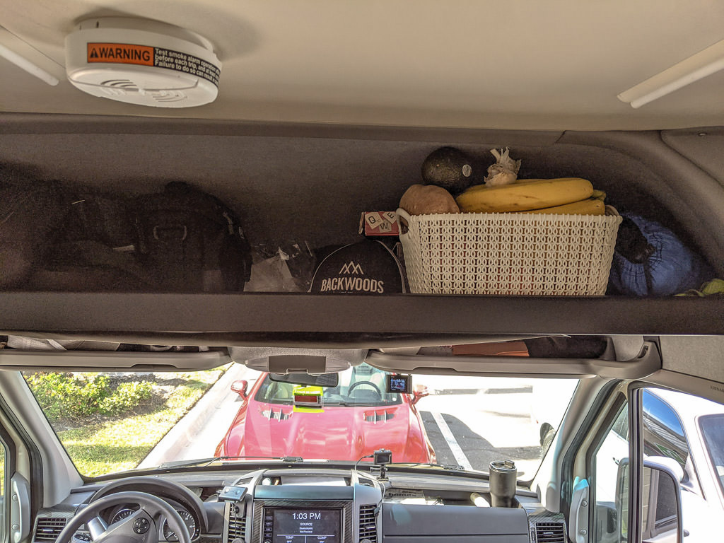 Overhead shelf above cab with white basket full of fruit, a baseball cap, a game and other supplies