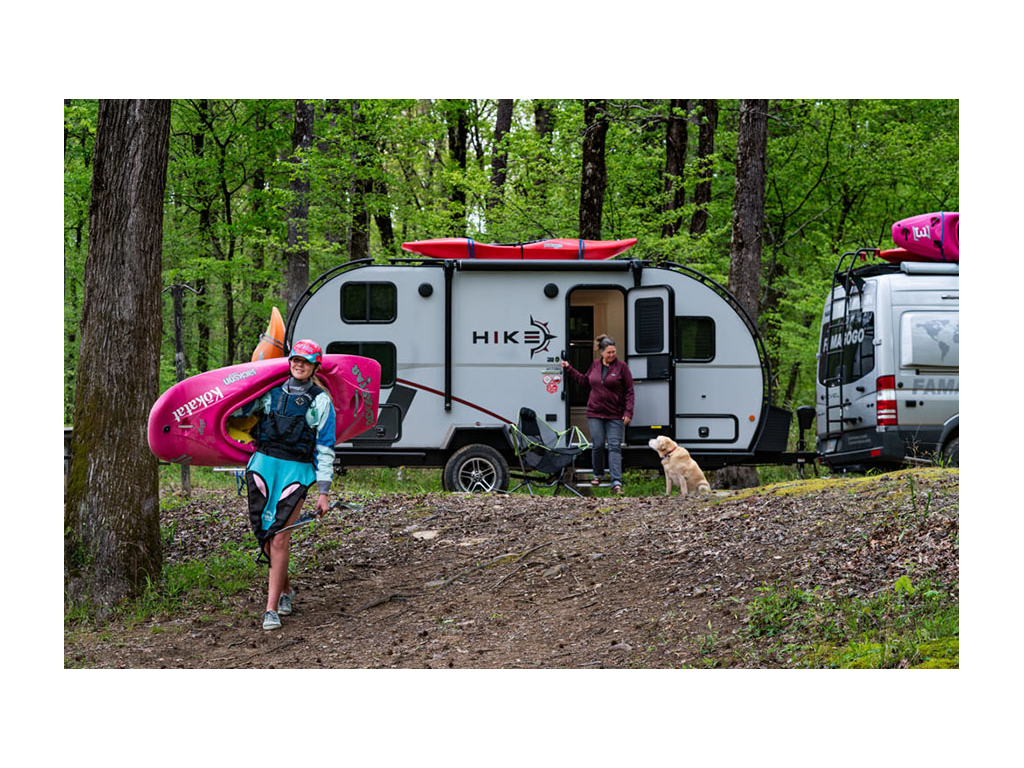Kathy standing at door of HIKE with yellow lab, Tucker, and Abby walking towards camera carrying red kayak