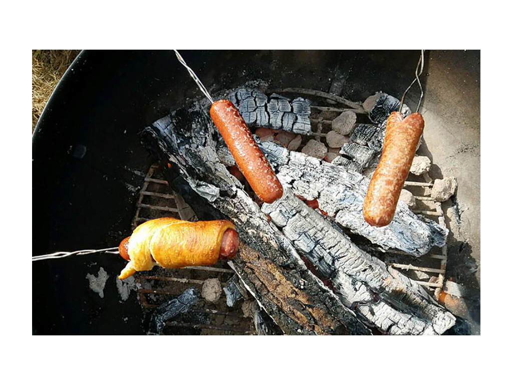 Three hot dogs on sticks roasting over campfire. One hot dog is wrapped in a crescent roll