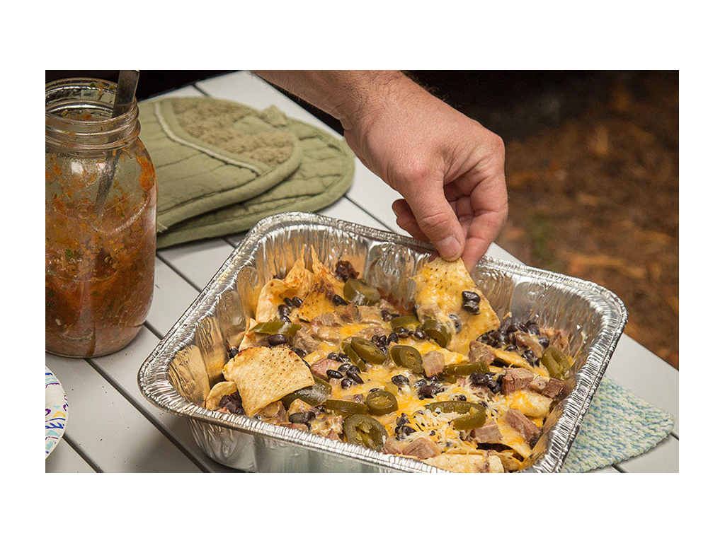A table with nachos in a tin pan, a jar of salsa, and green oven mitts
