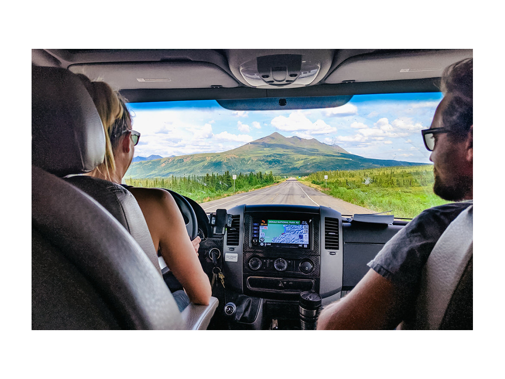 Photo from behind Katelyn driving with Howard in passenger seat .Looking out windshield to open road and mountains