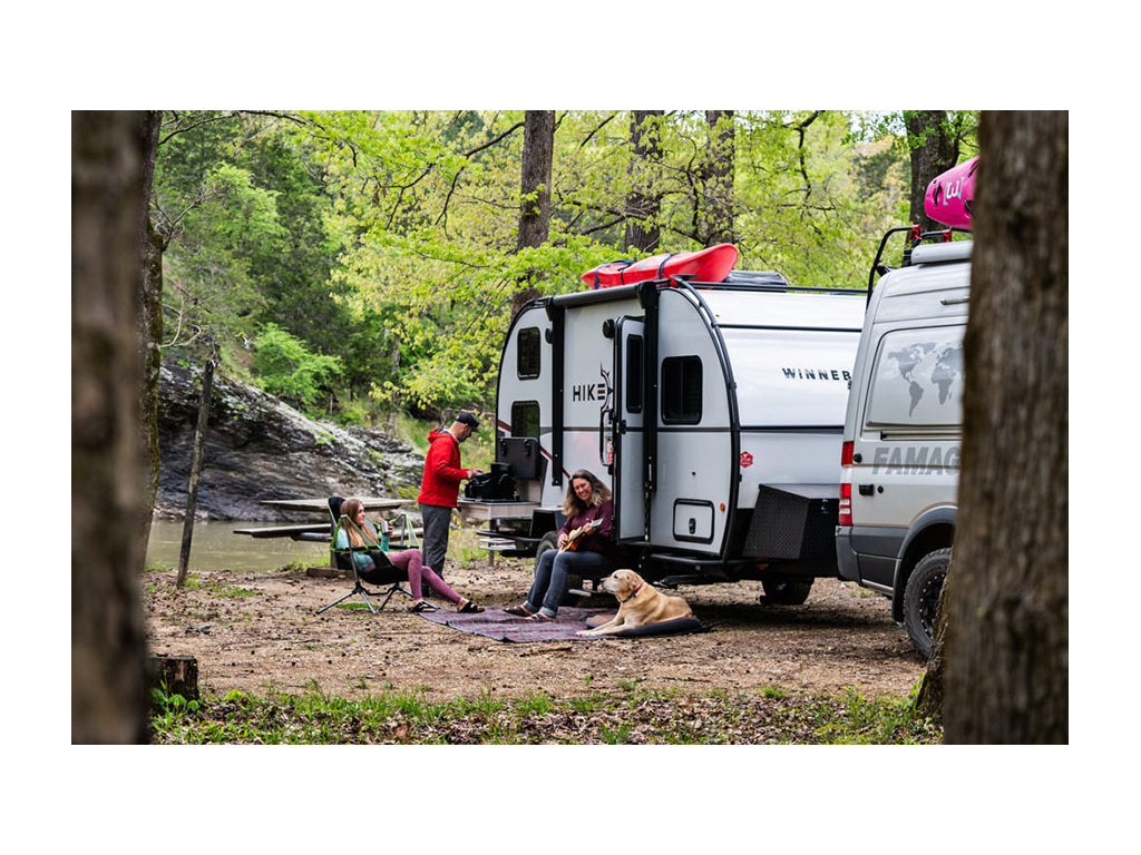 Kathy sitting on steps of HIKE playing ukulele next to yellow lab, Tucker. Peter making breakfast at outdoor kitchen while Abby sits in camp chair.