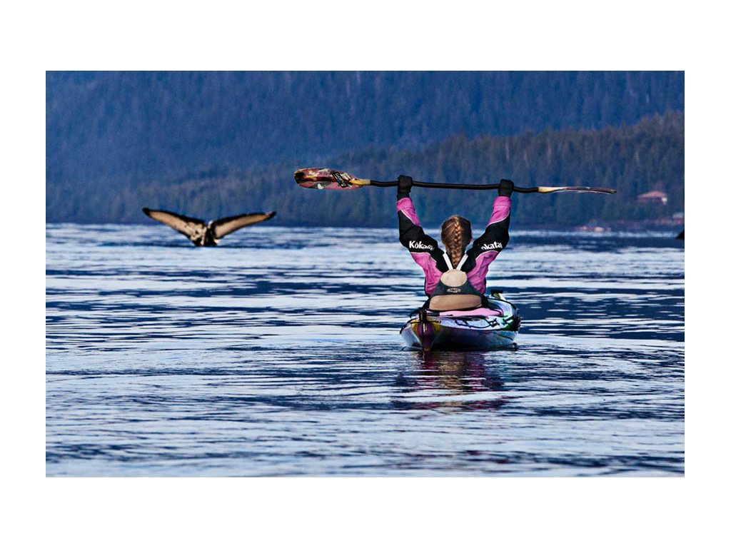Abby kayaking and holding up paddle with large bird flying nearby