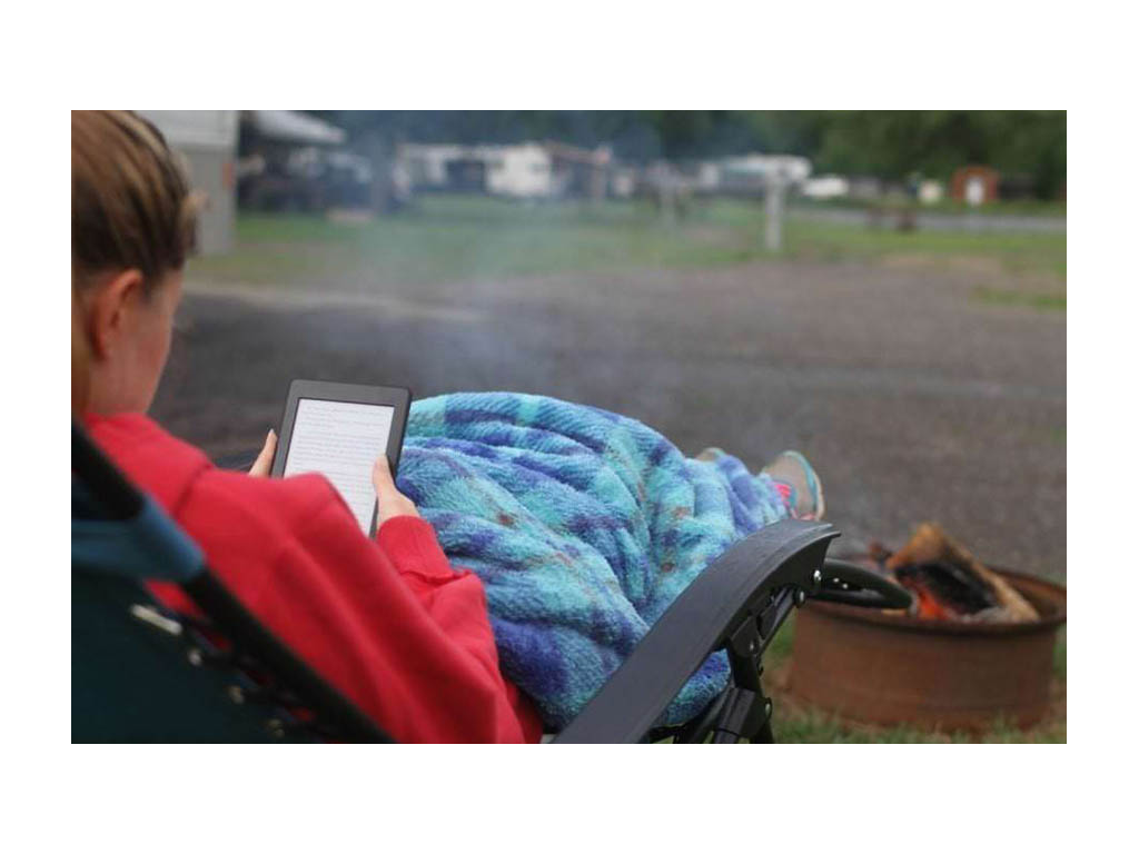 Alyssa sitting outside in camp chair wrapped in a blanket reading on a Kindle