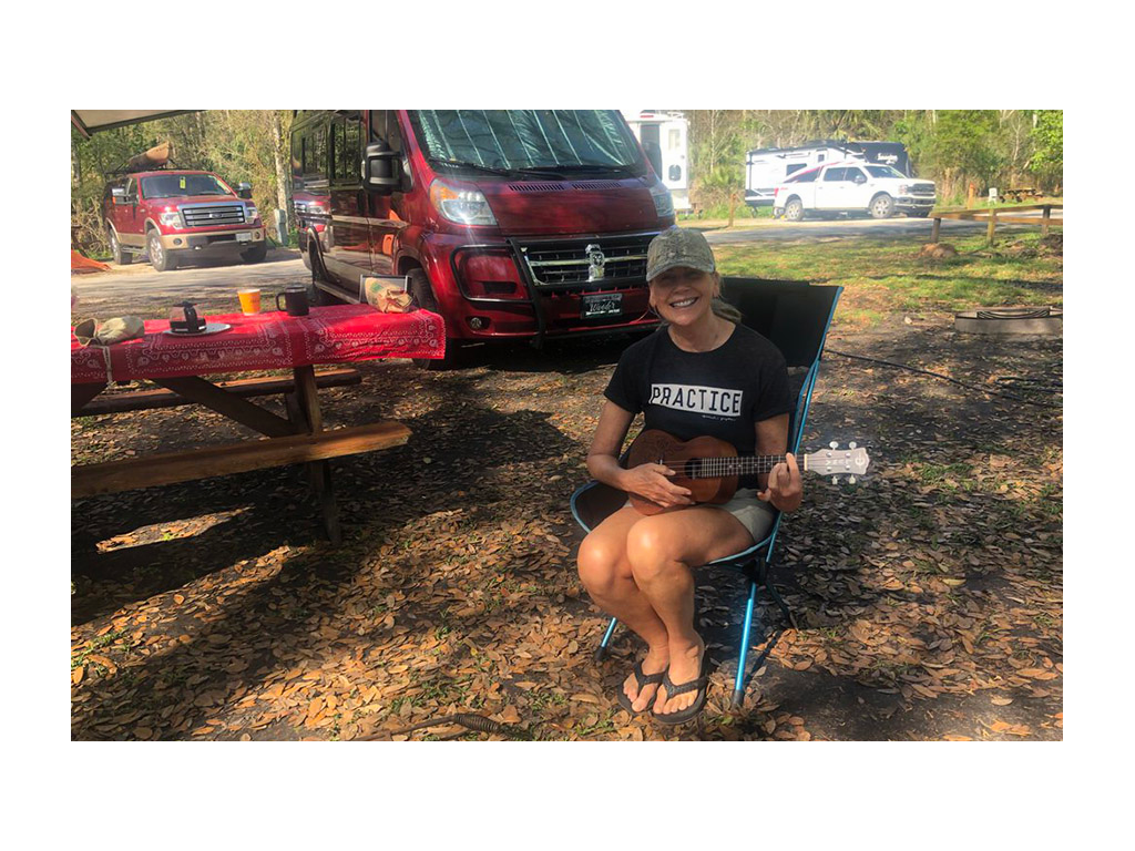 Viki playing ukulele in camp chair next to Travato at campground. 