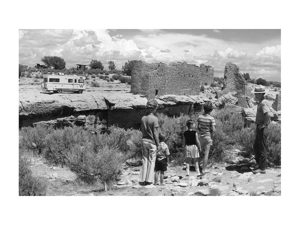 Black and white photo of park ranger leading a family of four through park. Old Winnebago Brave in the distance