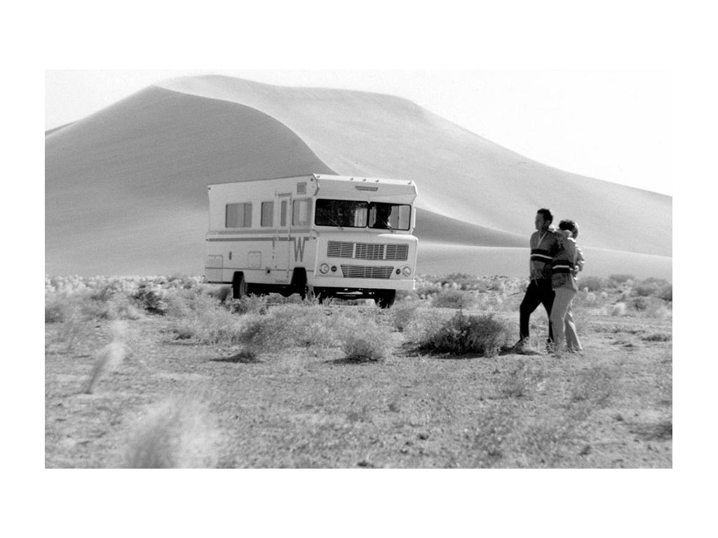Black and white photo of two people and a Winnebago in the sand dunes