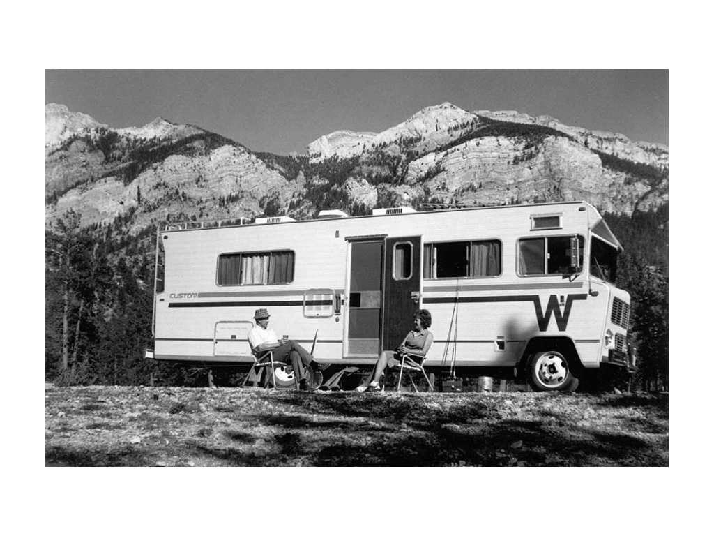 Old black and white photo of a couple sitting outside their Winnebago with mountains in the background