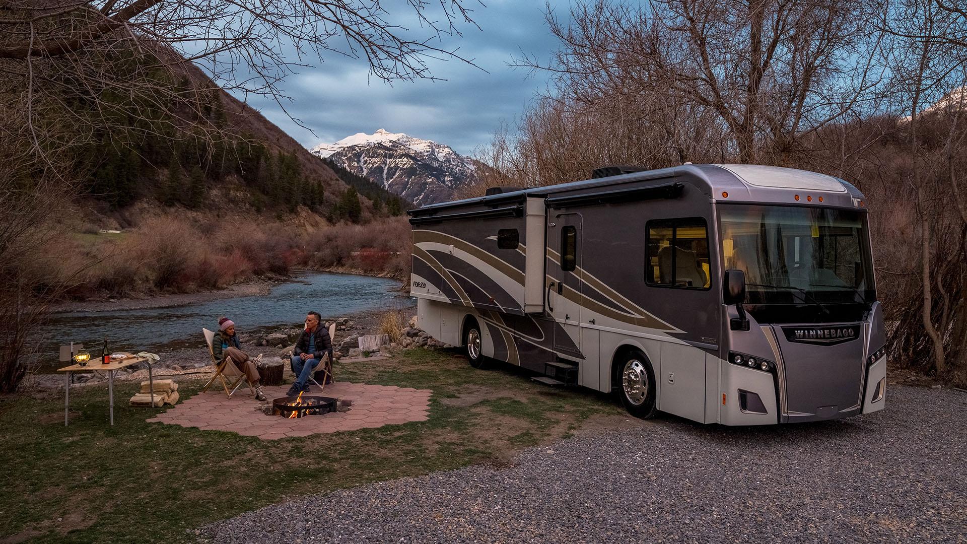 Two people sitting by a campfire beside their Winnebago RV, with a body of water and mountain in the background