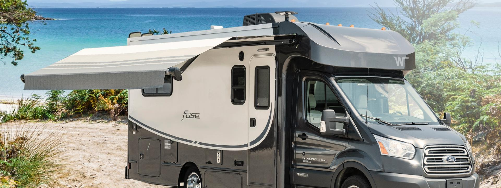 Winnebago Fuse exterior view with awning extended, on a sandy dune with water in the background