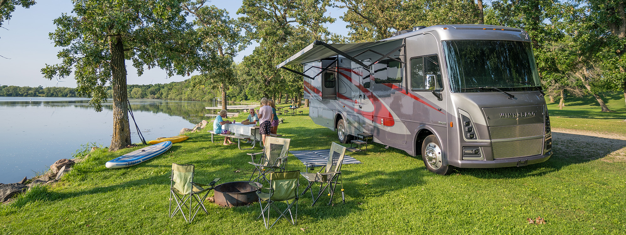 Winnebago Vista parked beside a body of water, with a group of people gathered around a nearby table