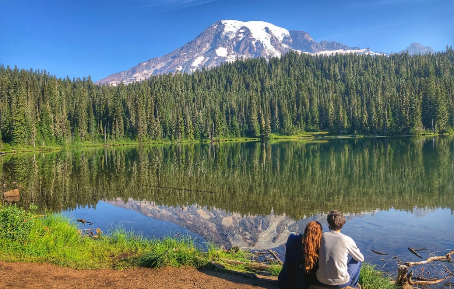 Couple gazing out over body of water, lined with evergreens and mountains in view.