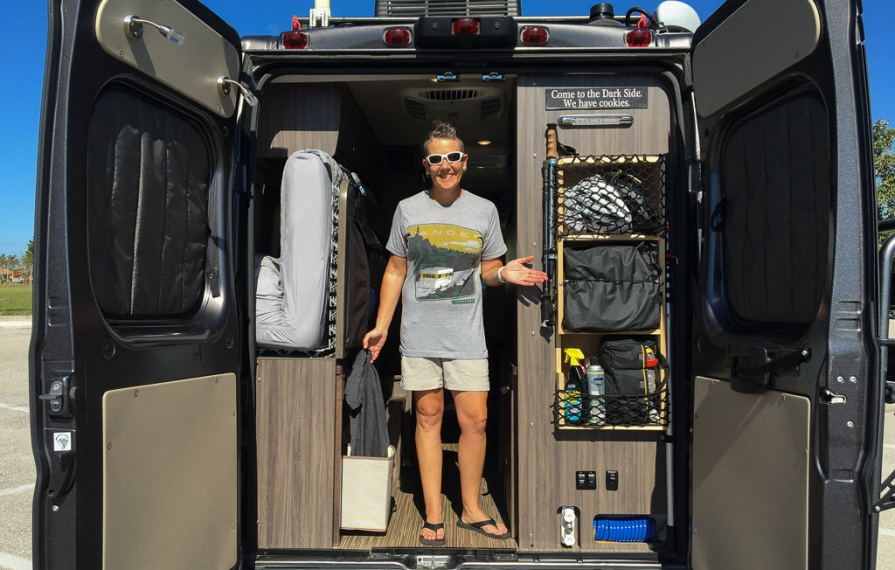 Noel standing in the back of the Travato with the rear doors open showing their modification of added storage