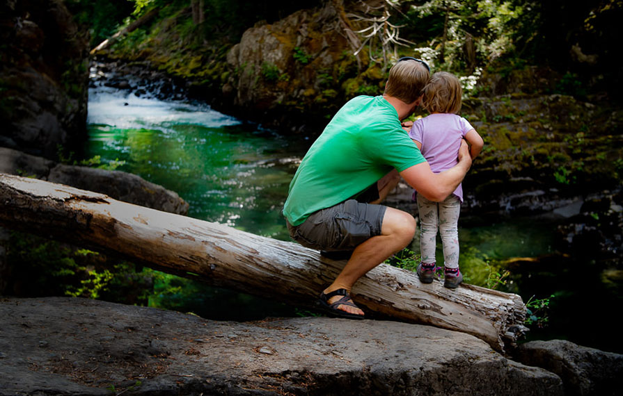 Dad and daughter sitting on a log looking out over a flowing stream.