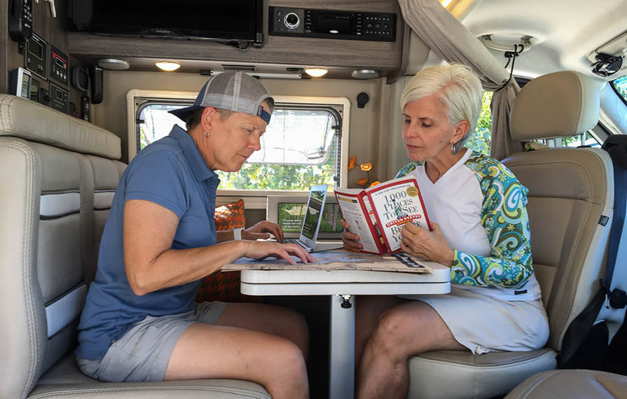 Noel and Chris sitting at dinette planning travels. Noel is looking at a map with laptop open and Chris is reading a travel destination book