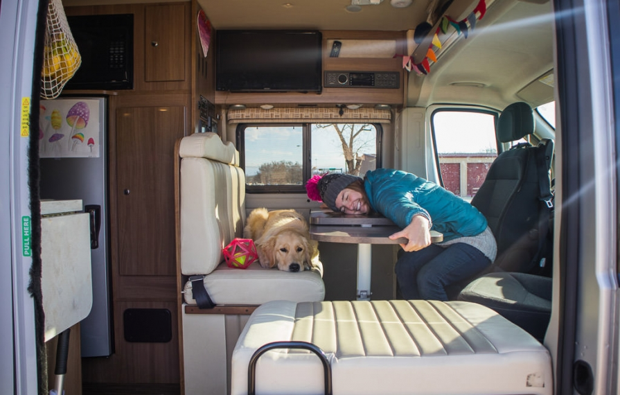 Woman laying head on table behind the driver's seat and dog on the bench of Winnebago Travato.