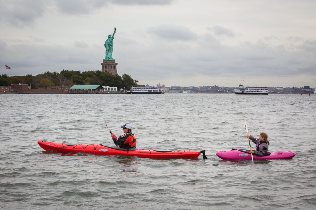 Two ladies kayaking on the water with the Statue of Liberty in the background.