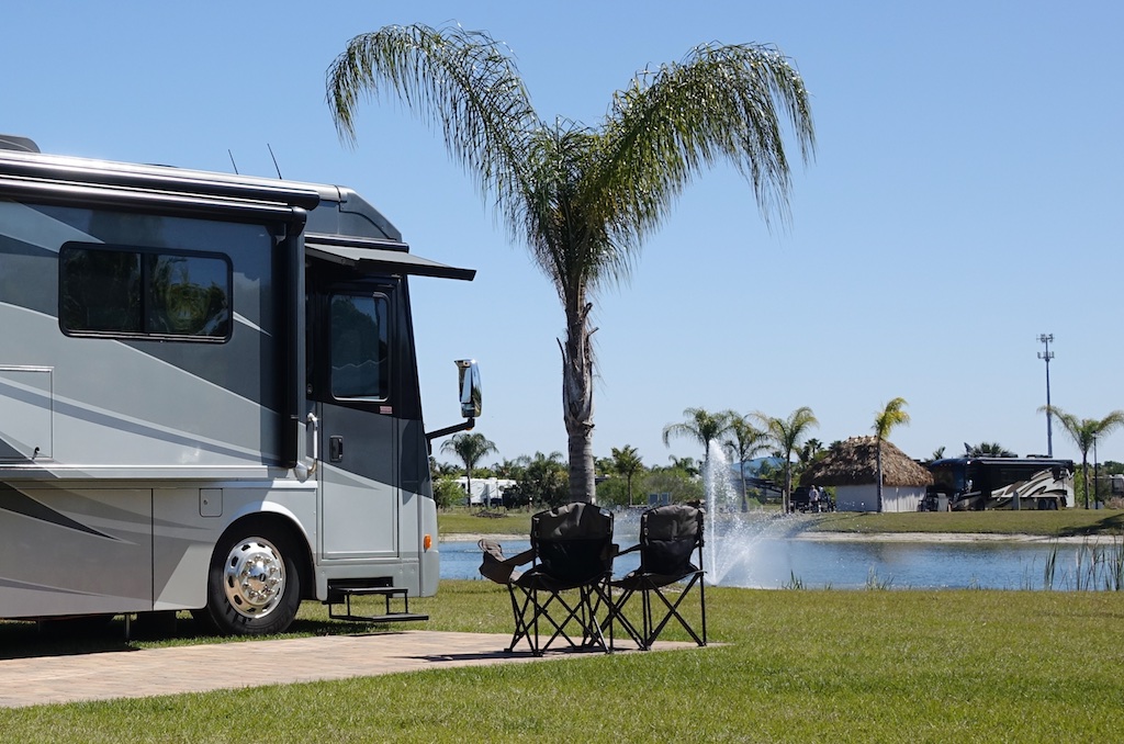 Two camping chairs sitting outside a motorhome with palm tree at site and water with fountain in front.