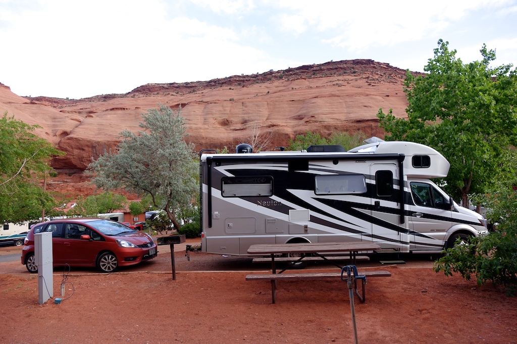 Winnebago Navion and car parked at campsite with red sandstone cliffs in the background.