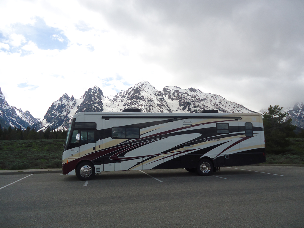 Winnebago Adventurer parked in lot with the snow covered Rocky Mountains behind.