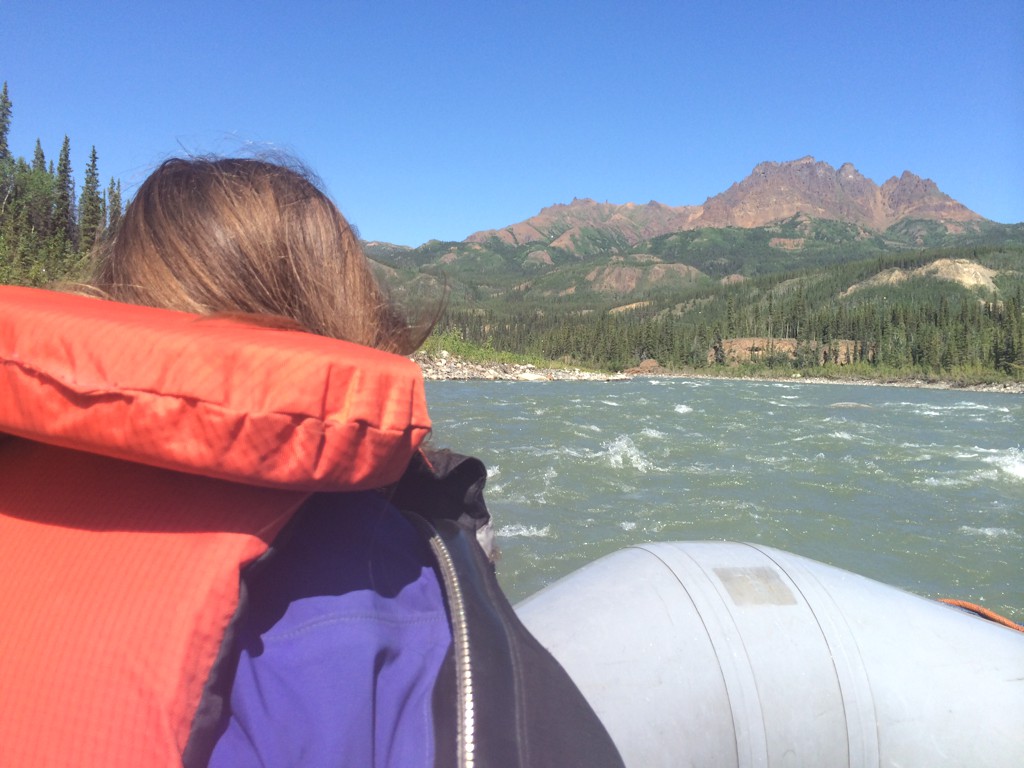 Alyssa at the front of a raft going down the river with mountains ahead.