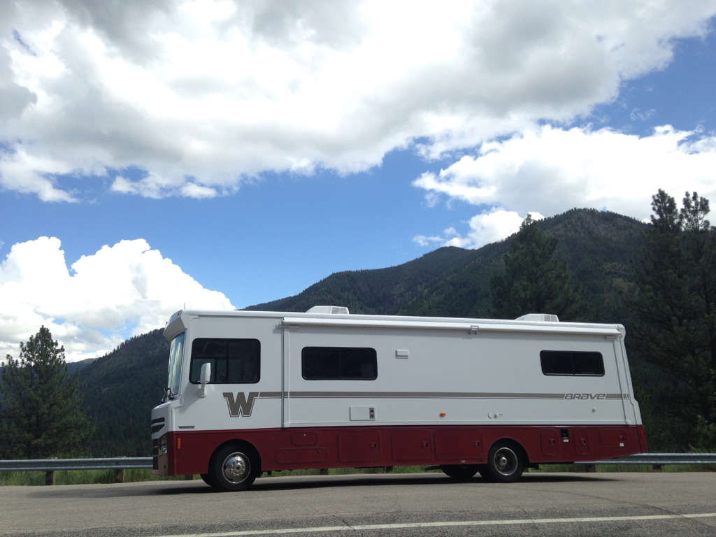 Winnebago Brave parked on the side of the road with tree covered high hills in the background.