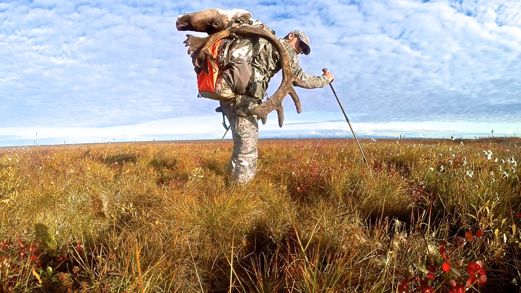 Damon with Caribou head strapped on his back while standing in grassy field.