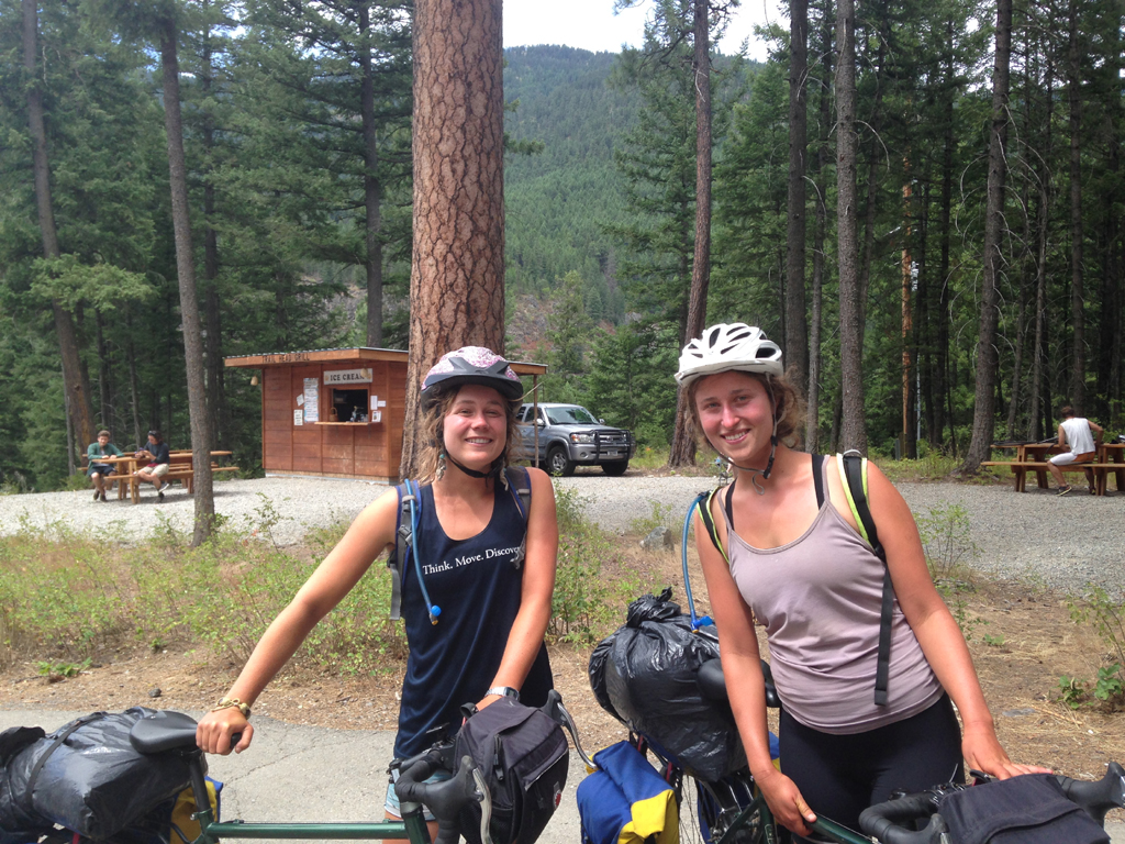 Two women with their bikes smiling at the camera with tall trees behind.