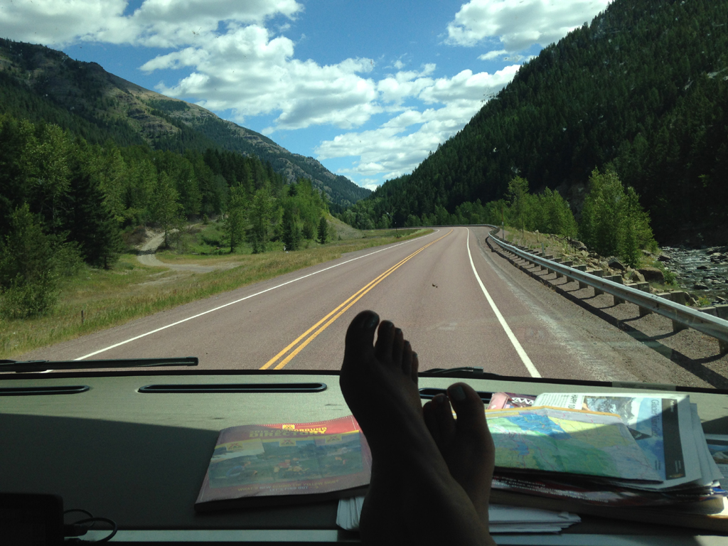 Feet resting on dash of vehicle driving down a road winding through tree covered hillsides.