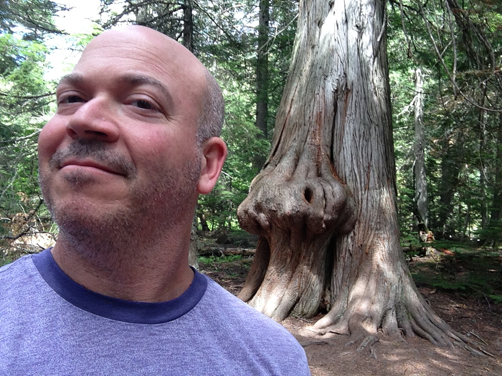 Man posing next to tree that has a growth that resembles a nose.