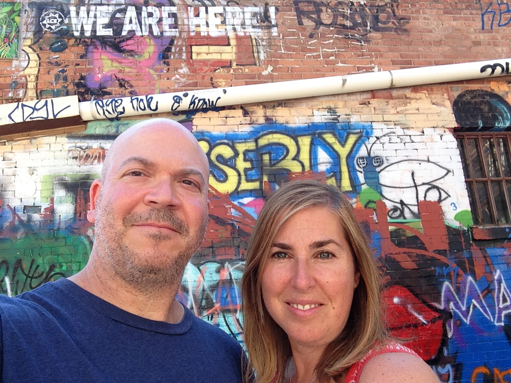 Couple taking a selfie with graffiti covered brick wall behind them. 