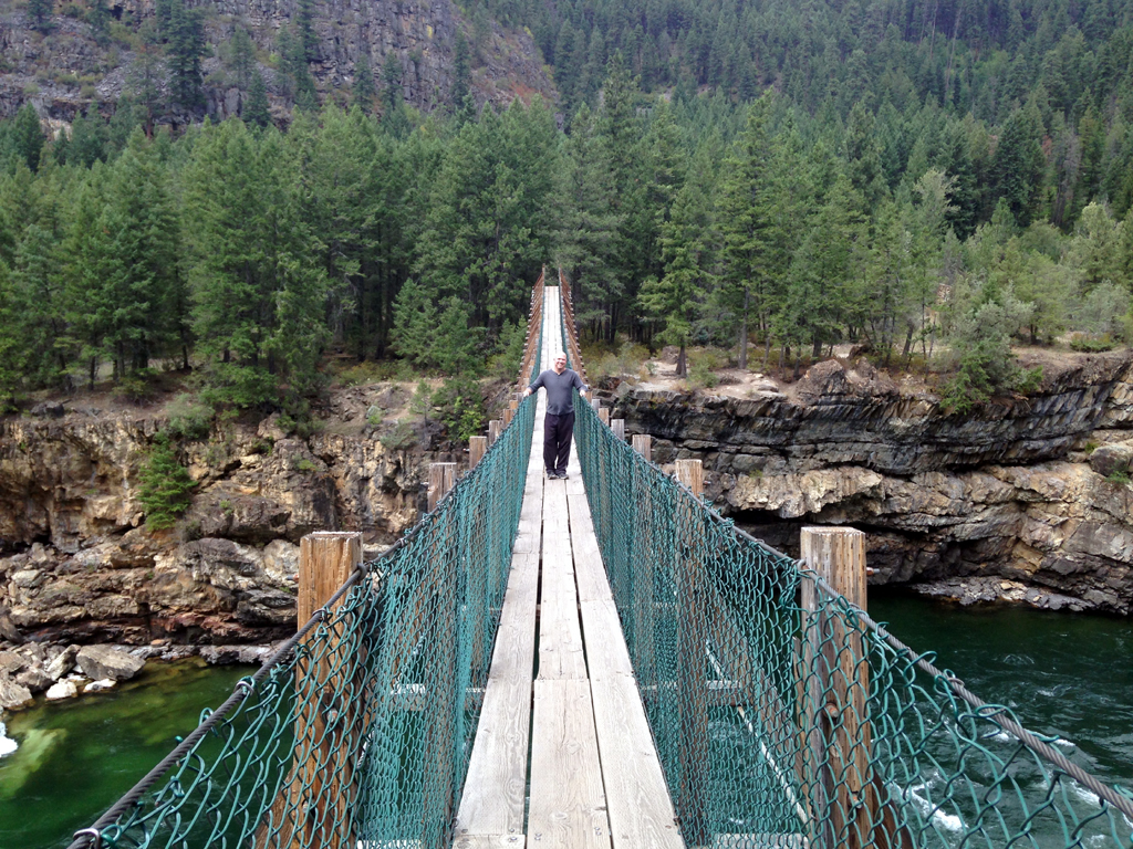 Man on swinging bridge over water with tree covered hillside behind him.