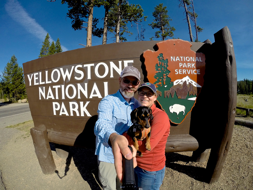 Couple with a dog taking a selfie in front of Yellowstone National Park sign.