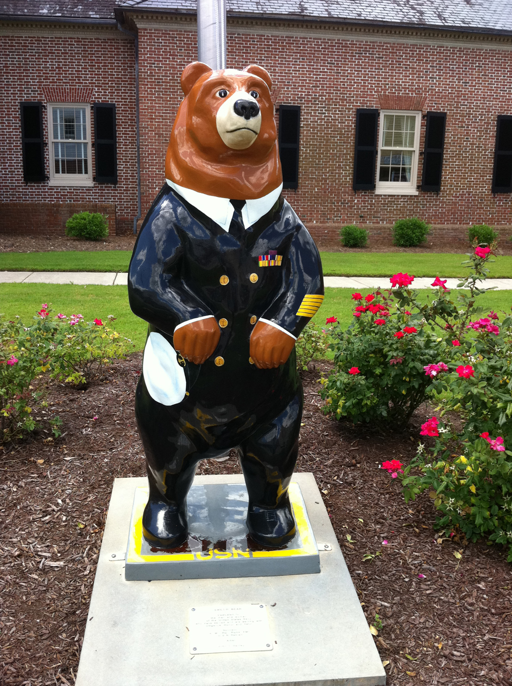 Life-size, hand painted fiberglass bear dressed in Navy blues in flower garden in front of a building.