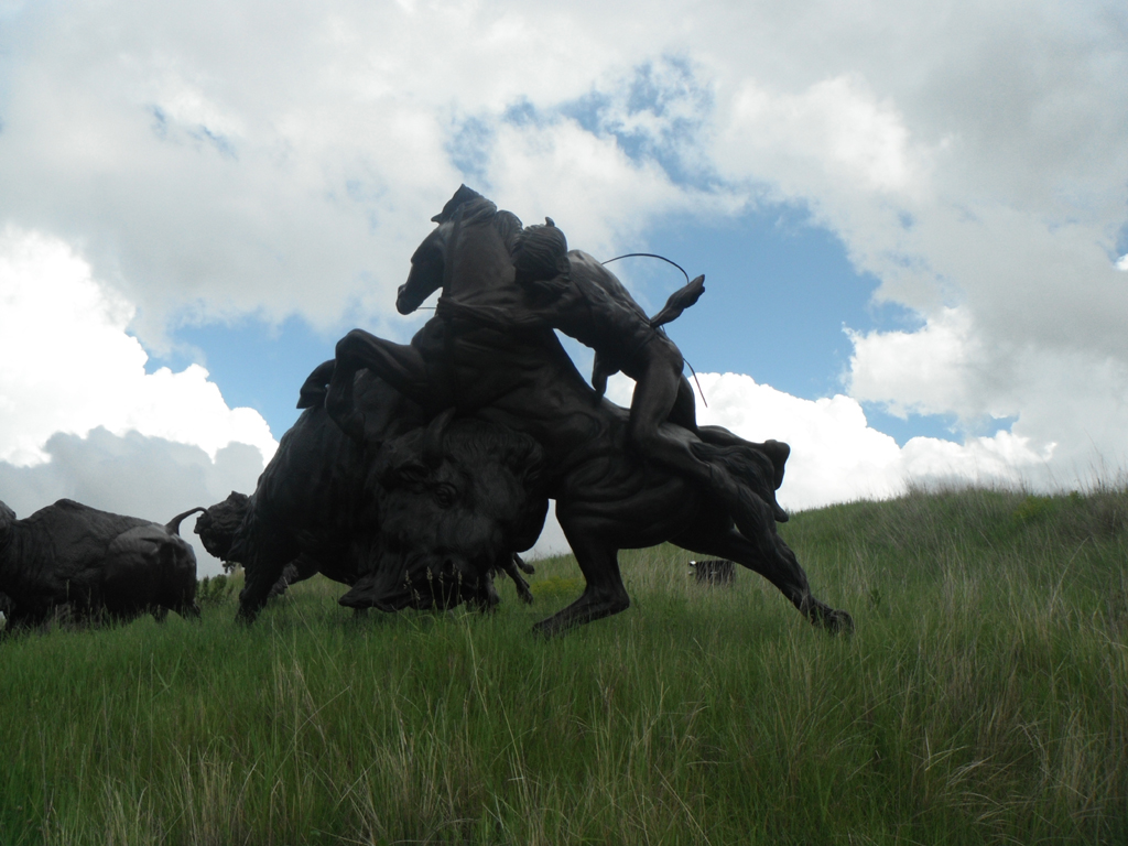 Sculpture in grassy field of Native American on horseback pursuing bison.