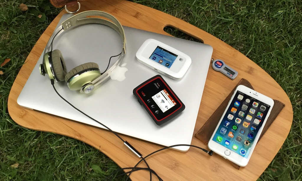 Headphones, 2 hotspot devices and an Iphone sitting on a lap desk on the grass.