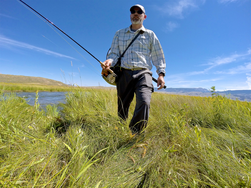 Man walking through grass with a fishing pole next to water.