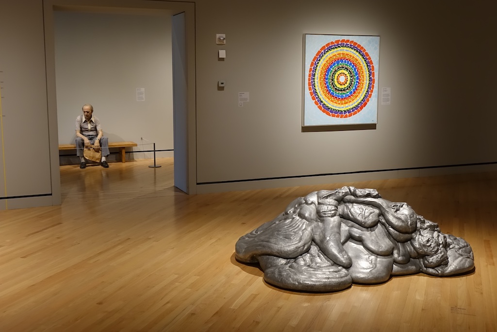 Sculpture on floor, painting on wall, and life-like sculpture of man sitting on a bench.