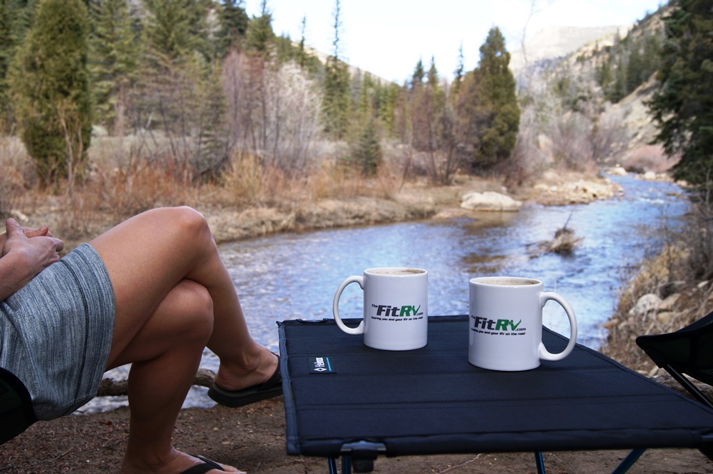 Coffee mugs that read FIT RV sitting on a camping table sitting at the edge of a river.