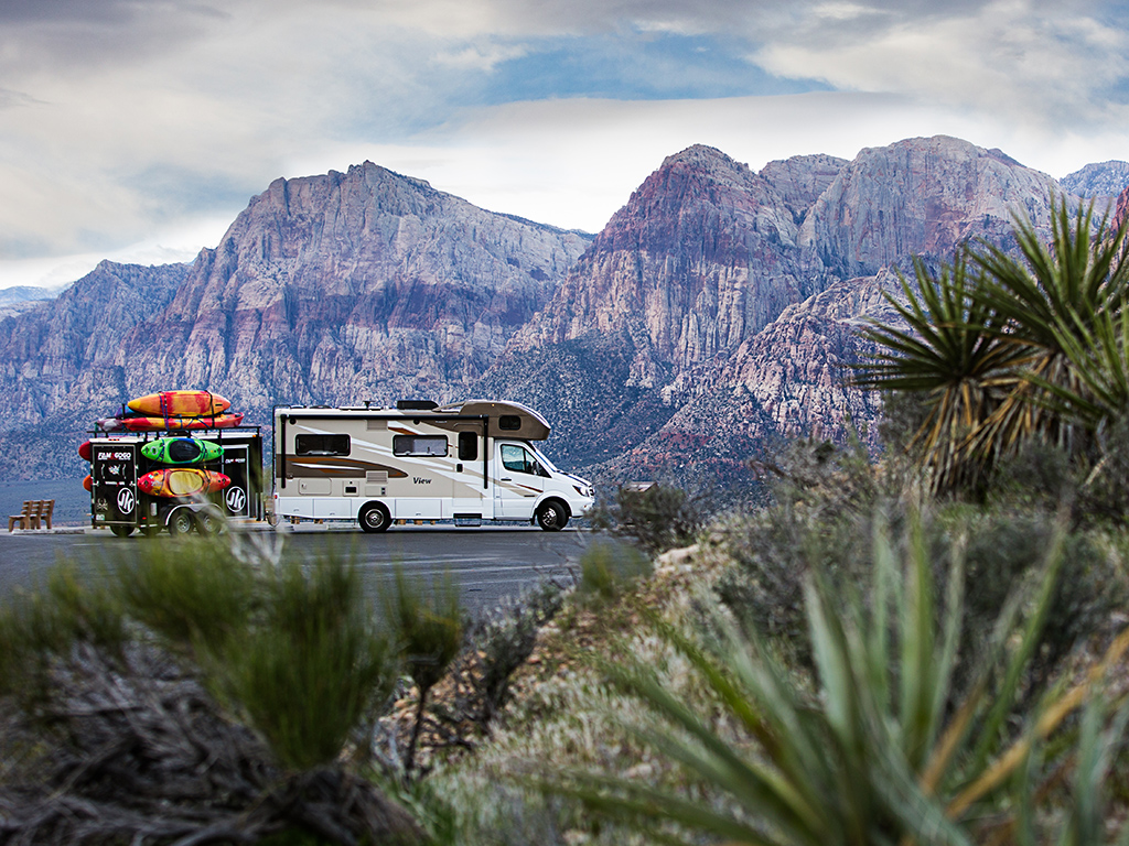 Winnebago View with trailer attached parked in lot with mountains behind. 